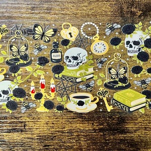 Può includere: Un foglio di adesivi decorativi con illustrazioni a tema gotico. Il design include teschi, farfalle, tazze da tè, libri, chiavi e orologi, in una palette di nero, giallo e bianco. Lo sfondo è un legno con venature.