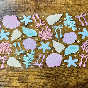 Puede incluir: Una hoja de pegatinas con diseños de temática marina. Las pegatinas incluyen conchas marinas, estrellas de mar, coral, algas marinas y lazos en colores pastel como rosa, azul y amarillo. Pequeñas estrellas salpican el diseño.