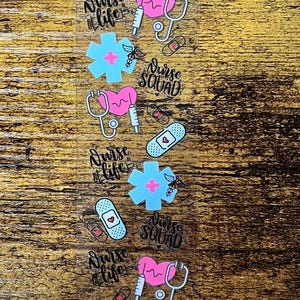 May include: Clear sticker sheet with medical-themed designs. Includes pink hearts, stethoscopes, syringes, bandages, and blue crosses. Text reads "Nurse Life" and "Nurse Squad" in a script font. The stickers are arranged on a wooden background.