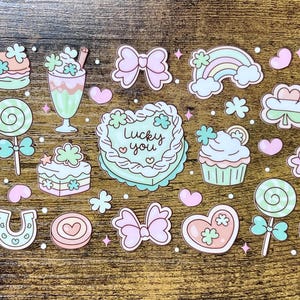 Op de afbeelding: Een vel stickers met een thema voor St. Patrick's Day. De stickers bevatten klavers, strikken, regenbogen, harten, cupcakes en een taart met de tekst "Lucky you". Het kleurenpalet is pastelroze, groen en blauw.