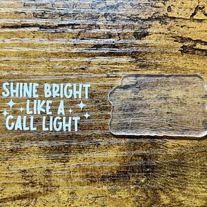 Könnte beinhalten: Ein klarer Acryl-Form und ein weißer Aufkleber mit dem Text "SHINE BRIGHT LIKE A CALL LIGHT" und Stern-Grafiken. Die Artikel befinden sich auf einer Holzoberfläche mit Holzmaserung.