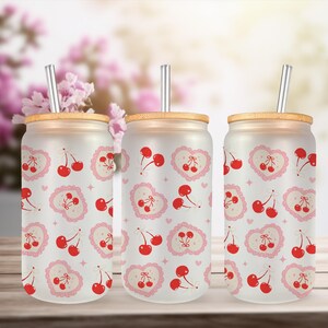 May include: Three frosted glass tumblers with bamboo lids and metal straws. Each tumbler features a repeating pattern of red cherries and pink heart-shaped designs. The background includes blurred pink flowers.