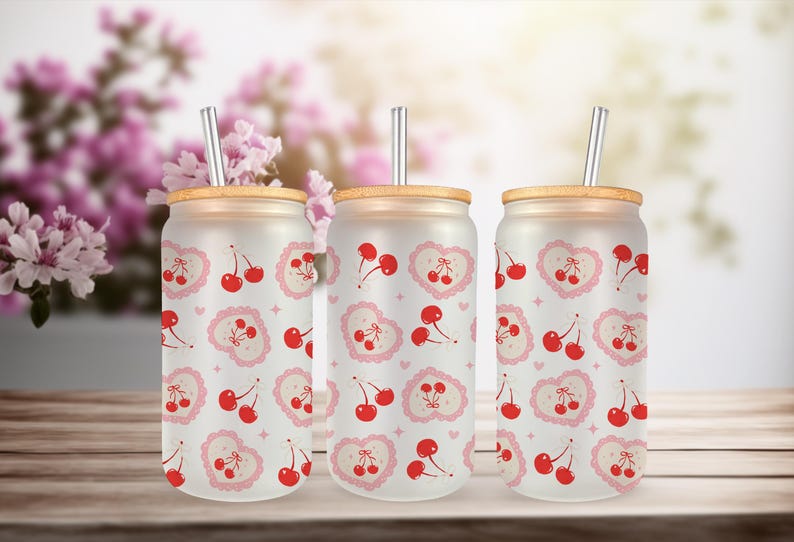 May include: Three frosted glass tumblers with bamboo lids and metal straws. Each tumbler features a repeating pattern of red cherries and pink heart-shaped designs. The background includes blurred pink flowers.