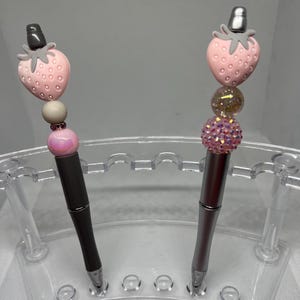 May include: Two silver pens with pink and gray strawberry-shaped toppers and decorative beads. The pens are displayed in a clear plastic stand.