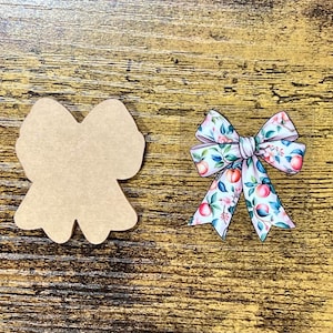May include: Two bow-shaped ornaments. One is a blank, light brown shape. The other is a colorful bow with a floral pattern of peaches, green leaves, and pink flowers on a white background. The bow has a white center.