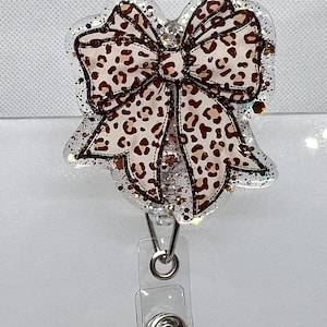 May include: A clear plastic badge holder with a brown and white leopard print bow. The bow has gold glitter accents.