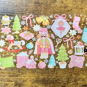 Puede incluir: Una colección de pegatinas con temática navideña que presenta cascanueces, árboles de Navidad, adornos y una bailarina. Las pegatinas son de colores pastel rosa, verde y azul, con detalles dorados. Dispuestas sobre una superficie de madera.