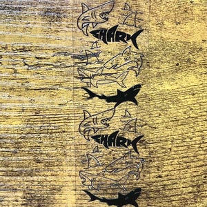 May include: A clear, rectangular sticker featuring a repeating pattern of shark illustrations. The word "SHARK" is incorporated into the design. The sharks are depicted in black, with a variety of poses and styles.
