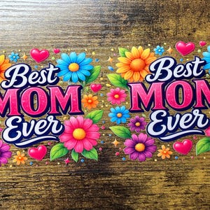 May include: Two colorful floral designs with the text "Best MOM Ever". The designs feature vibrant flowers in shades of pink, orange, blue, and purple, along with pink hearts and green leaves. The background is a wooden texture.