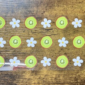 May include: A sheet of stickers featuring a repeating pattern of kiwi fruit slices and white flowers. The kiwi slices are green with a brown border and black seeds. The flowers have white petals and yellow centers.
