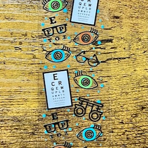 Puede incluir: Un marcapáginas transparente con ilustraciones relacionadas con los ojos. El diseño incluye gafas, tablas optométricas y ojos estilizados en verde azulado, naranja y verde. El texto "E FP TOZ" es visible. El marcapáginas está sobre una superficie de madera.