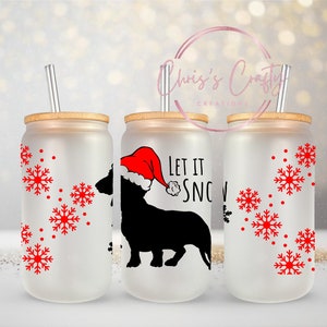 Puede incluir: Tres vasos de vidrio esmerilado con tapas de bambú y pajitas rojas. Los vasos tienen un fondo blanco con copos de nieve rojos y una silueta negra de un dachshund con un gorro de Papá Noel. Un vaso tiene el texto "Let it snow".