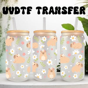 May include: Three glass drinkware with bamboo lids and straws. The drinkware features a capybara and floral pattern. The text "UVDTF TRANSFER" is at the top. The background includes greenery.