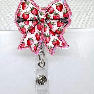 May include: A pink glitter badge reel with a white bow featuring a strawberry pattern. The reel has a clear plastic retractable cord and a silver clip.