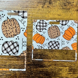 May include: Two rectangular badge holders with a fall theme. The holders feature a light blue background with orange pumpkins, brown pumpkins with black spots, and black and white checkered pumpkins. Black swirls and dots complete the design.