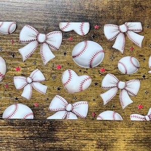 May include: A collection of baseball-themed stickers and embellishments. The image features baseballs, heart-shaped baseballs, and white bows with red stitching. Scattered red stars and black star shapes add to the design on a wood-grain background.