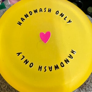 May include: A bright yellow, circular object with the words "HANDWASH ONLY" in black, curved around the edge. A small, pink heart sticker is centered on the object. The text is repeated on the bottom.