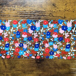 May include: A rectangular floral design featuring red, blue, and pink flowers with white stems and leaves. Small red bows and blue hearts are interspersed throughout the design. The background is a dark wood grain.