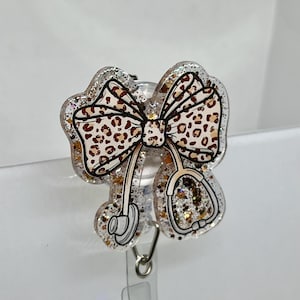 May include: A clear plastic badge holder with a gold glitter and leopard print bow and a stethoscope design. The holder has a retractable clip.