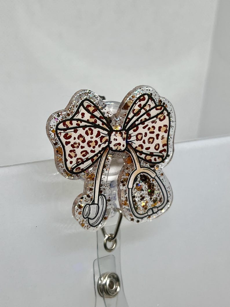 May include: A clear plastic badge holder with a gold glitter and leopard print bow and a stethoscope design. The holder has a retractable clip.