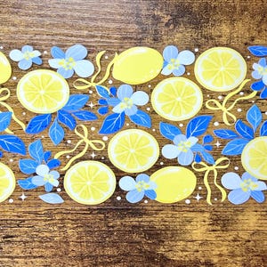 Könnte beinhalten: Dekoratives Design mit gelben Zitronen, Zitronenscheiben und blauen Blumen mit gelben Bändern. Die Anordnung befindet sich vor einem Holzgrund, wodurch eine fröhliche und sommerliche Ästhetik entsteht.