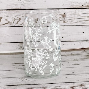 May include: A clear glass vase with a white floral design. The vase is cylindrical and has a wide mouth. The floral design is simple and elegant, featuring leaves, flowers, and stems.