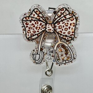 May include: A clear glitter badge reel with a leopard print bow and a stethoscope design. The reel has a silver clip and a silver ring.