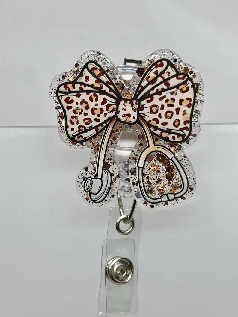 May include: A clear glitter badge reel with a leopard print bow and a stethoscope design. The reel has a silver clip and a silver ring.