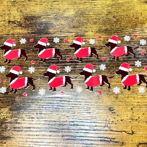 Puede incluir: Ocho pegatinas de perro salchicha negro y rojo con gorros de Papá Noel y sosteniendo un corazón rojo. Las pegatinas están sobre un fondo de madera.