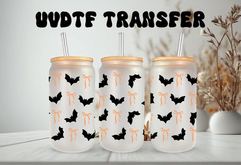 May include: Three frosted glass tumblers with bamboo lids and straws. Each tumbler is decorated with a pattern of black bats and orange bows. The text "UVDTF TRANSFER" is above the tumblers. Halloween-themed drinkware.