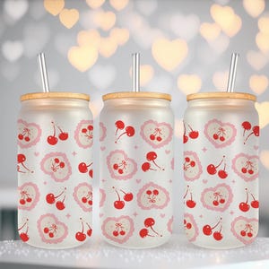 May include: Three frosted glass drink tumblers with bamboo lids and silver straws. Each tumbler features a repeating pattern of red cherries and pink hearts. The tumblers are designed for cold beverages.