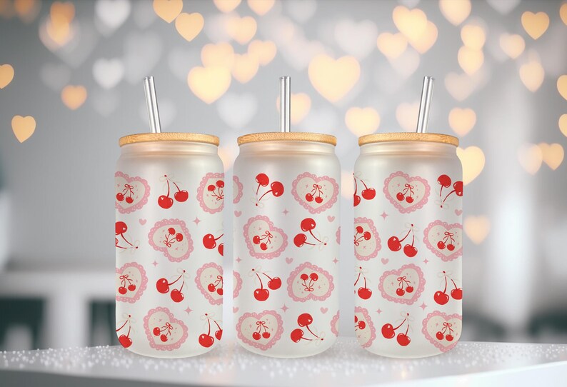 May include: Three frosted glass drink tumblers with bamboo lids and silver straws. Each tumbler features a repeating pattern of red cherries and pink hearts. The tumblers are designed for cold beverages.