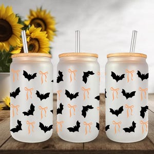 May include: Three frosted glass tumblers with bamboo lids and clear straws. Each tumbler features a repeating pattern of black bats and orange bows. The background includes sunflowers in a white vase, set on a wooden table.