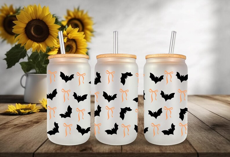 May include: Three frosted glass tumblers with bamboo lids and clear straws. Each tumbler features a repeating pattern of black bats and orange bows. The background includes sunflowers in a white vase, set on a wooden table.