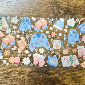 Puede incluir: Una hoja de pegatinas con ilustraciones de temática invernal. Los diseños incluyen una rebeca azul, mitones, una bufanda, un gorro, una tetera, una taza de chocolate caliente, libros, calcetines y copos de nieve. La paleta de colores es suave, con azules, rosas y blancos pastel.