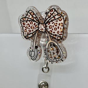 May include: A clear glitter badge holder with a brown and white leopard print bow and a stethoscope design. The holder has a clear plastic clip.