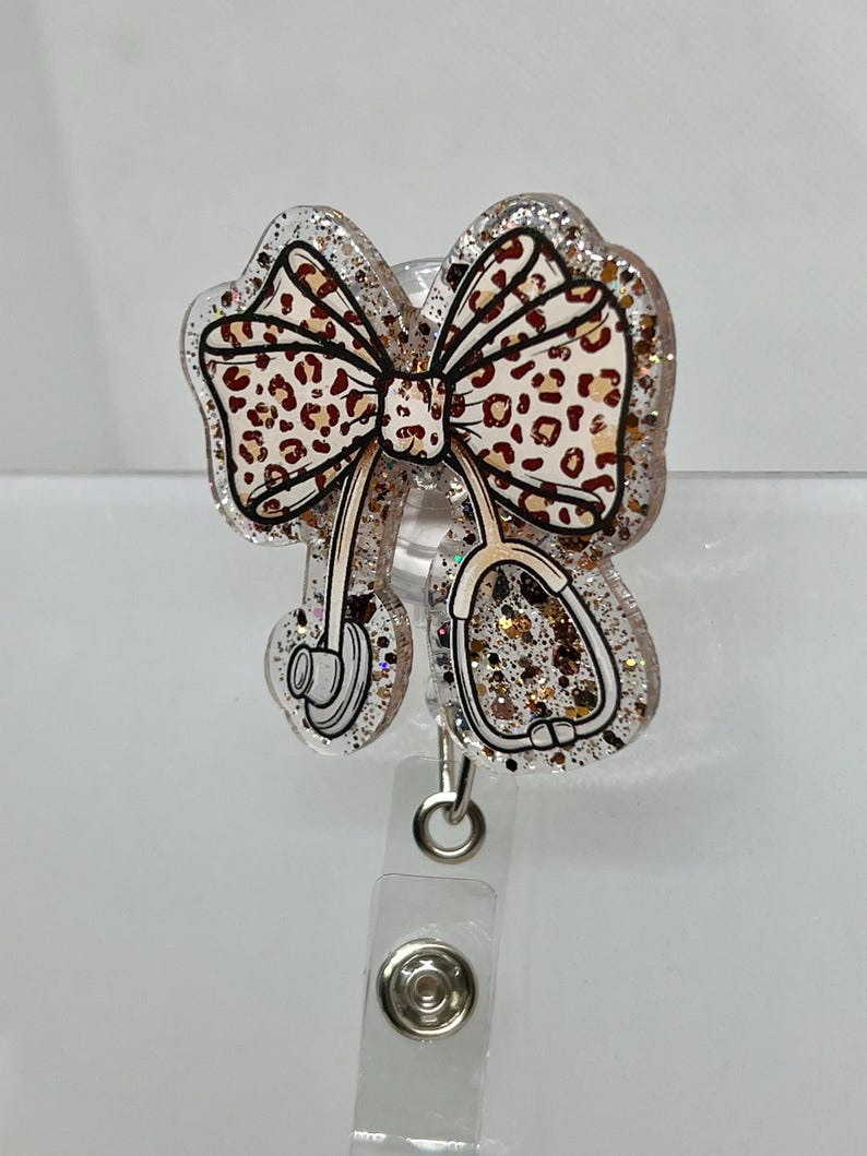 May include: A clear glitter badge holder with a brown and white leopard print bow and a stethoscope design. The holder has a clear plastic clip.