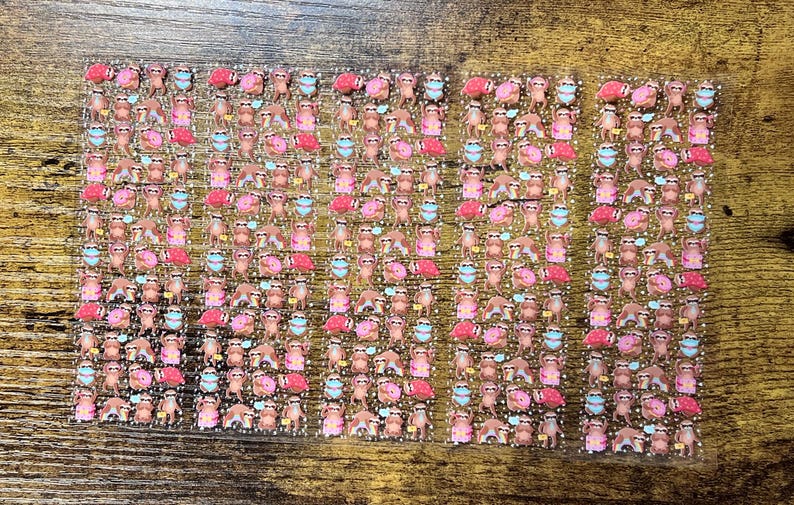 May include: Sheets of stickers featuring various sloth designs. The stickers showcase sloths in different poses, some wearing face masks, holding donuts, and with pink flamingos. The stickers are in shades of brown, pink, and blue.