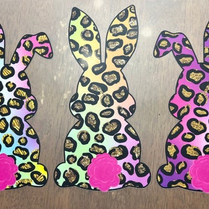 May include: Three pastel-colored Easter bunny cutouts with a leopard print pattern and gold glitter accents. Each bunny has a pink flower on its bottom.