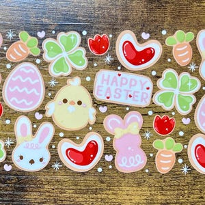 May include: A collection of Easter-themed stickers featuring various designs. Includes pink bunny and egg shapes, carrots, clovers, red heart-shaped cookies, and a yellow chick. The stickers have a white border and the text "HAPPY EASTER".