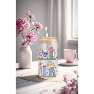 May include: A frosted glass tumbler with a wooden lid and straw, decorated with a whimsical bookshelf design. The design features books, a potion bottle, a rose, a sword, and a crown. A vase of pink flowers and a pink mug are in the background.