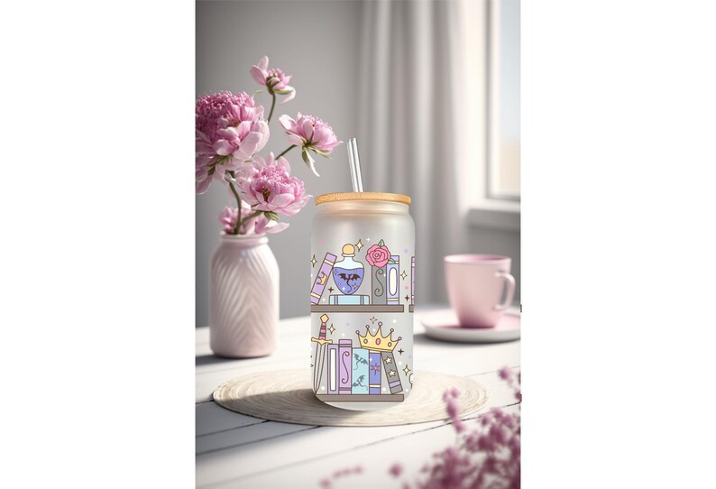 May include: A frosted glass tumbler with a wooden lid and straw, decorated with a whimsical bookshelf design. The design features books, a potion bottle, a rose, a sword, and a crown. A vase of pink flowers and a pink mug are in the background.