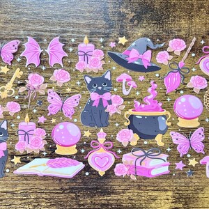 May include: A clear sticker sheet featuring various illustrations in shades of pink, purple, and black. Designs include black cats with pink bows, purple cauldrons, crystal balls, witch hats, butterflies, roses, books, potions, and small stars.