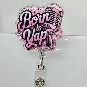 Pode incluir: Porta-crachá de resina rosa e preta com glitter, com um microfone e o texto "Born to Yap".