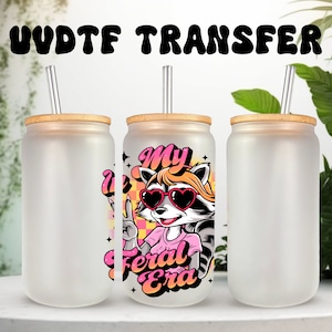 May include: Three frosted glass tumblers with bamboo lids and clear straws. The centre tumbler has a graphic of a cartoon raccoon wearing sunglasses and a pink shirt with the text "My Feral Era".
