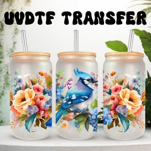 May include: Three frosted glass tumblers with lids and straws, featuring a blue jay and floral designs. The tumblers are on a white surface with green foliage in the background. The text "UV DTF TRANSFER" is at the top of the image.