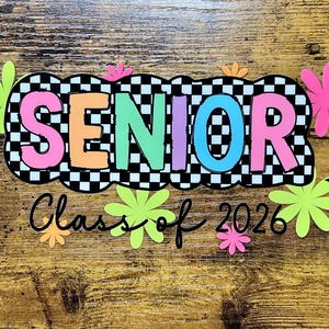 Può includere: Un colorato cartello "SENIOR Classe del 2026" con uno sfondo a scacchi in bianco e nero. La parola "SENIOR" è in colori vivaci e il design include fiori e stelle. Il cartello è su una superficie di legno.