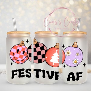 May include: Three frosted glass tumblers with bamboo lids and straws. Each tumbler features a different Christmas ornament design with a pink, black, white, and purple color scheme. The tumblers are arranged in a row and the text "FESTIVE AF" is printed on the front.
