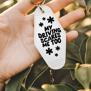 May include: White motel key chain with black text that reads "My driving scares me too". The keychain has a black flower design.
