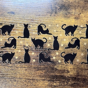 May include: A collection of black cat silhouettes in various poses, including sitting, stretching, and walking, interspersed with small gold-colored star shapes arranged across the surface in a repeating pattern.